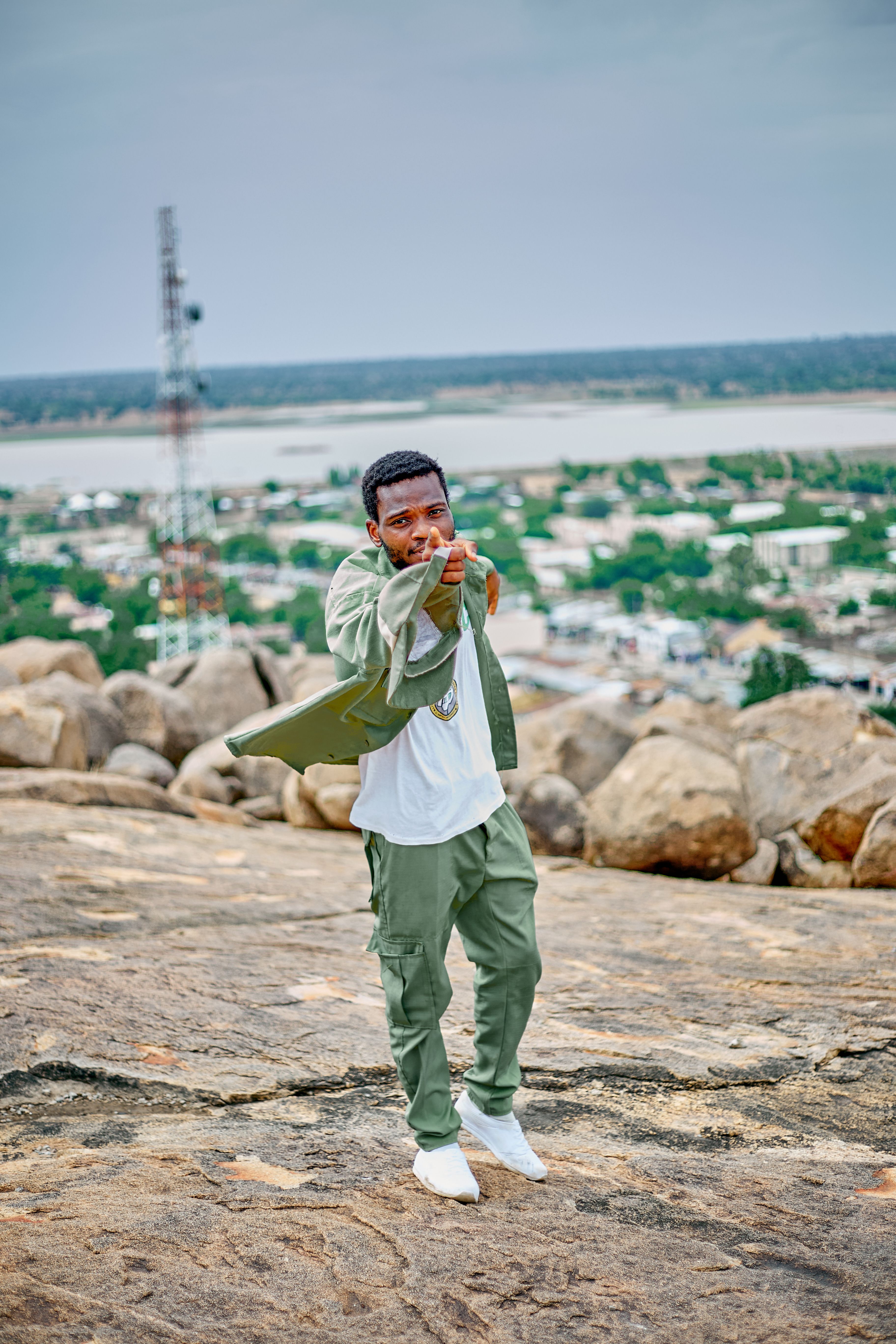 A young man poses confidently on a rocky hill with a cityscape in the background, showcasing urban style.