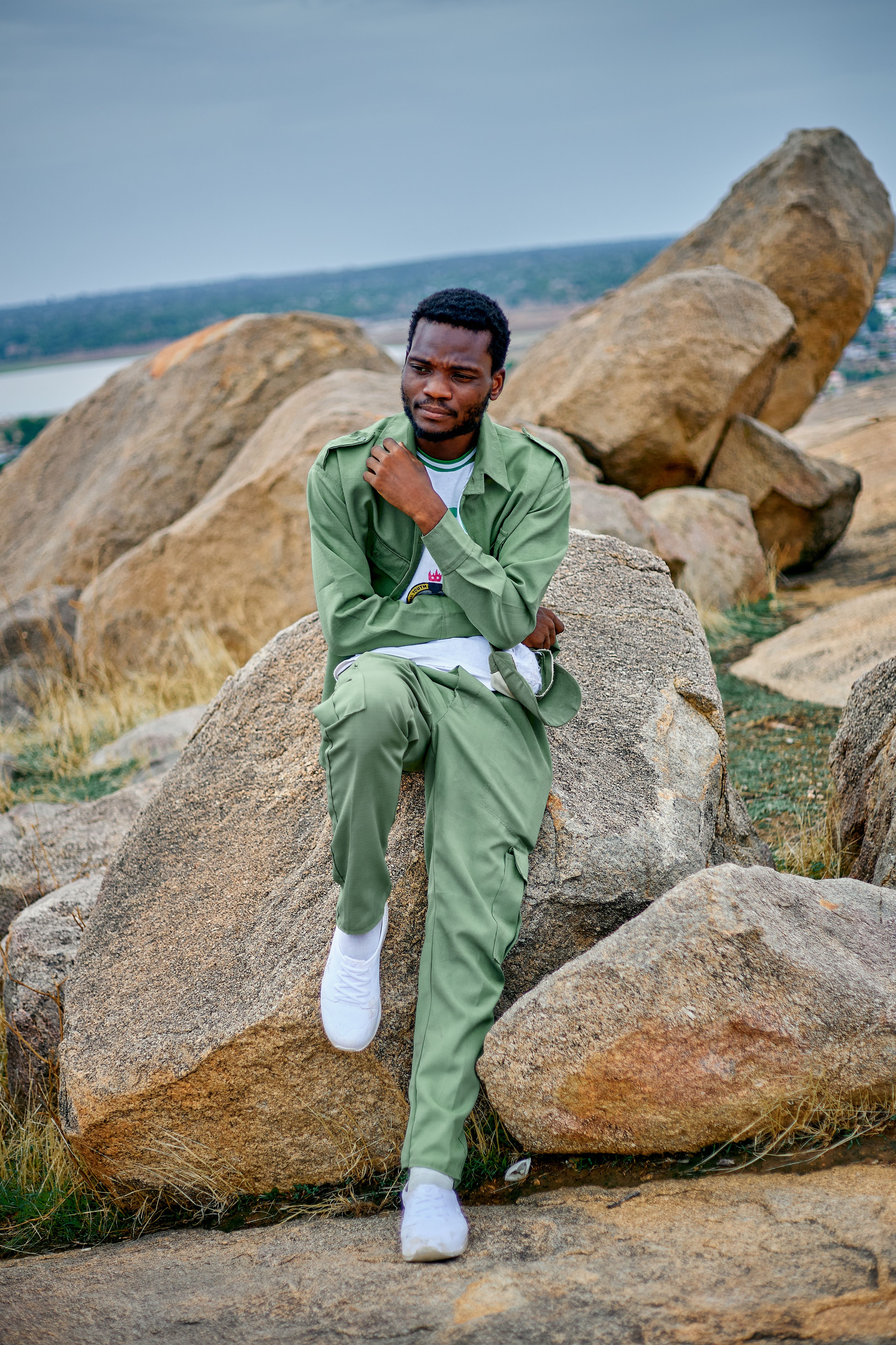 A man in a green suit sits thoughtfully on a rock outdoors, surrounded by nature.