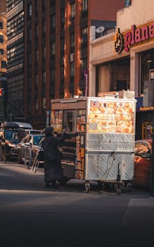 Street food cart in New York City at dusk. Urban scene with buildings and cars.