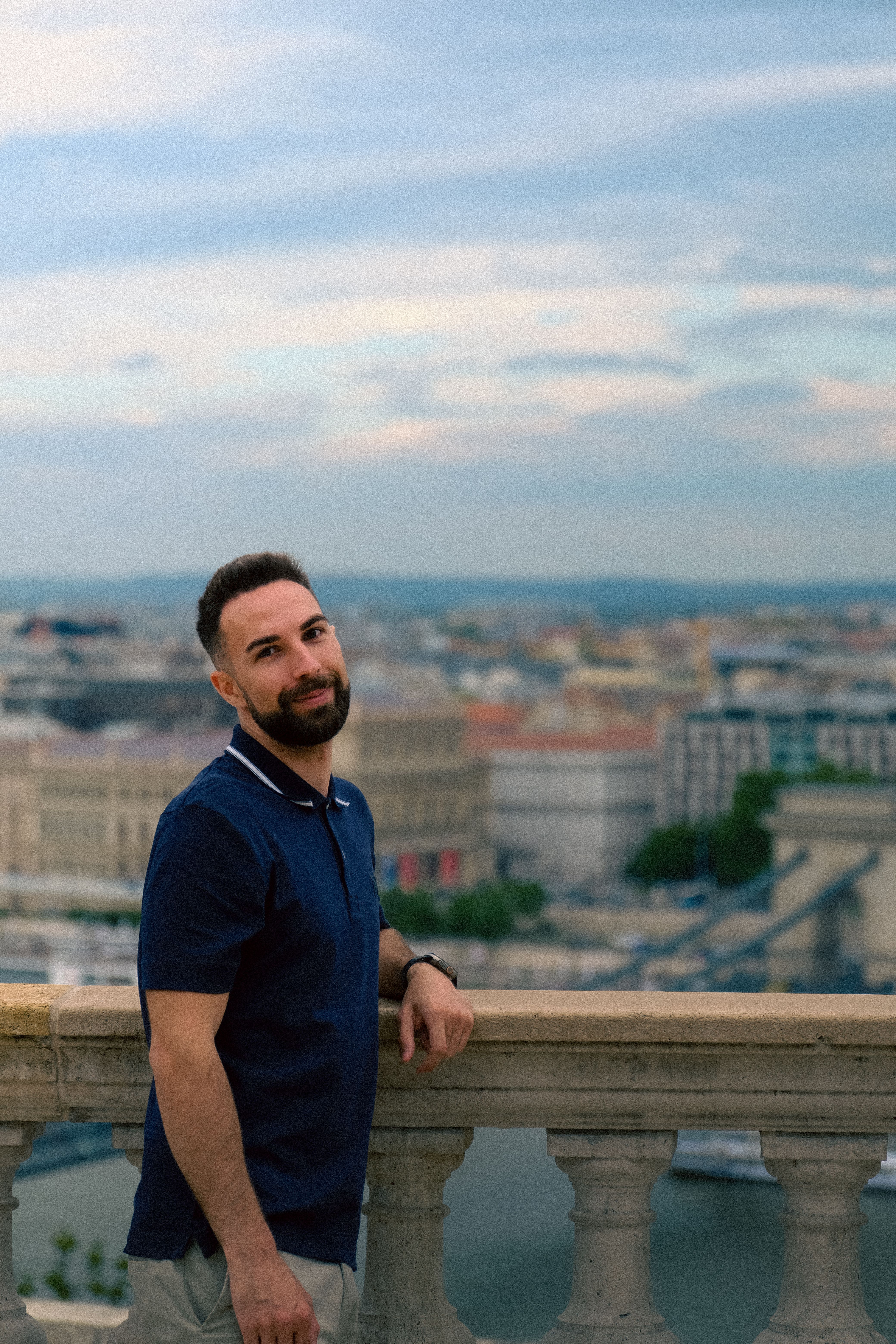 Young man poses on a balcony overlooking a scenic cityscape, captured during daytime.