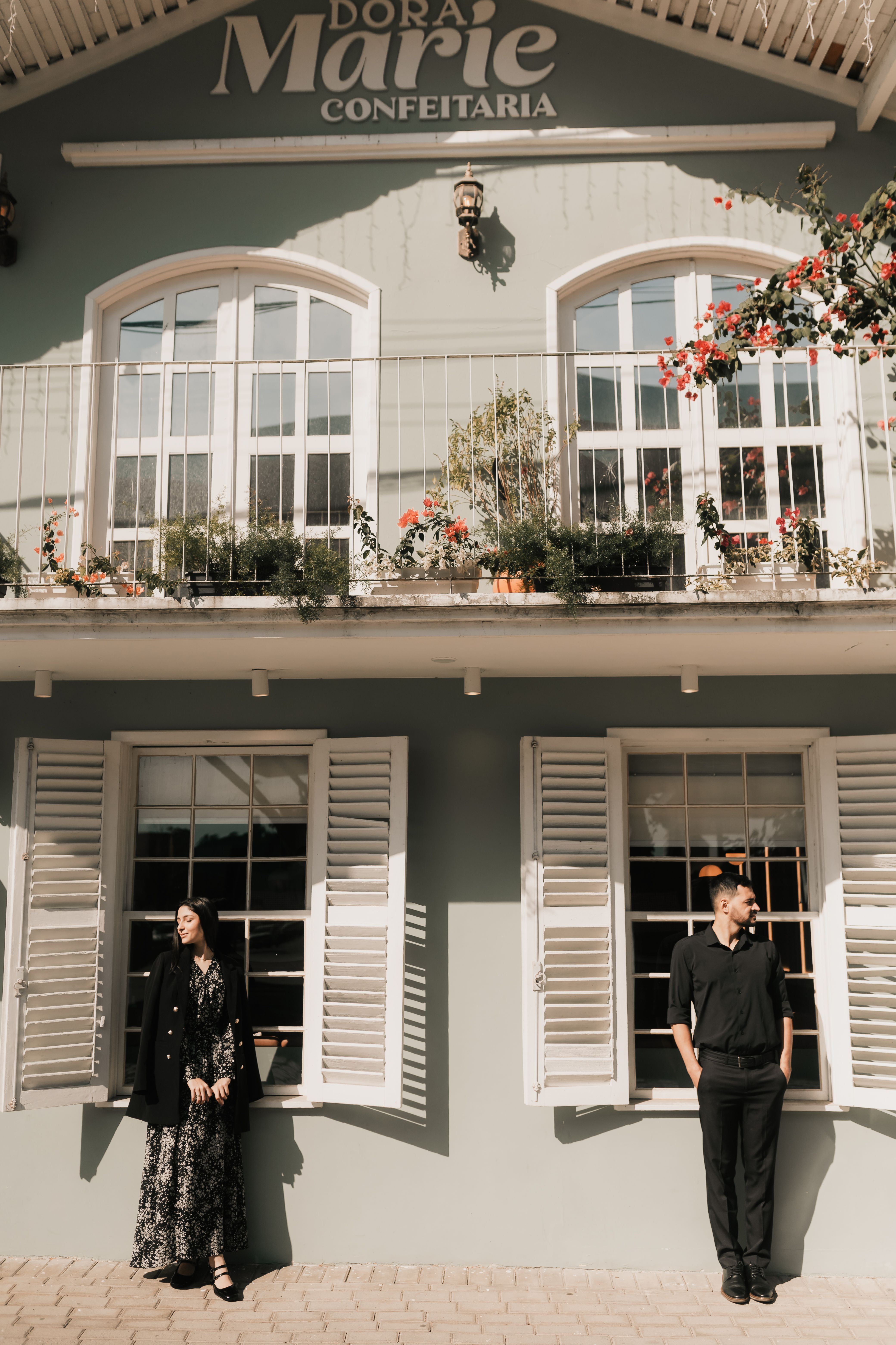 Stylish individuals posing by a quaint confectionery shop in a sunlit outdoor setting.