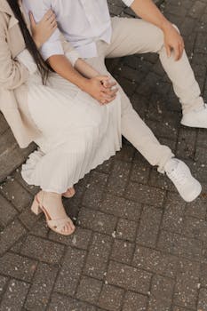 Stylish young couple sitting outdoors on paved ground wearing beige and white casual attire.