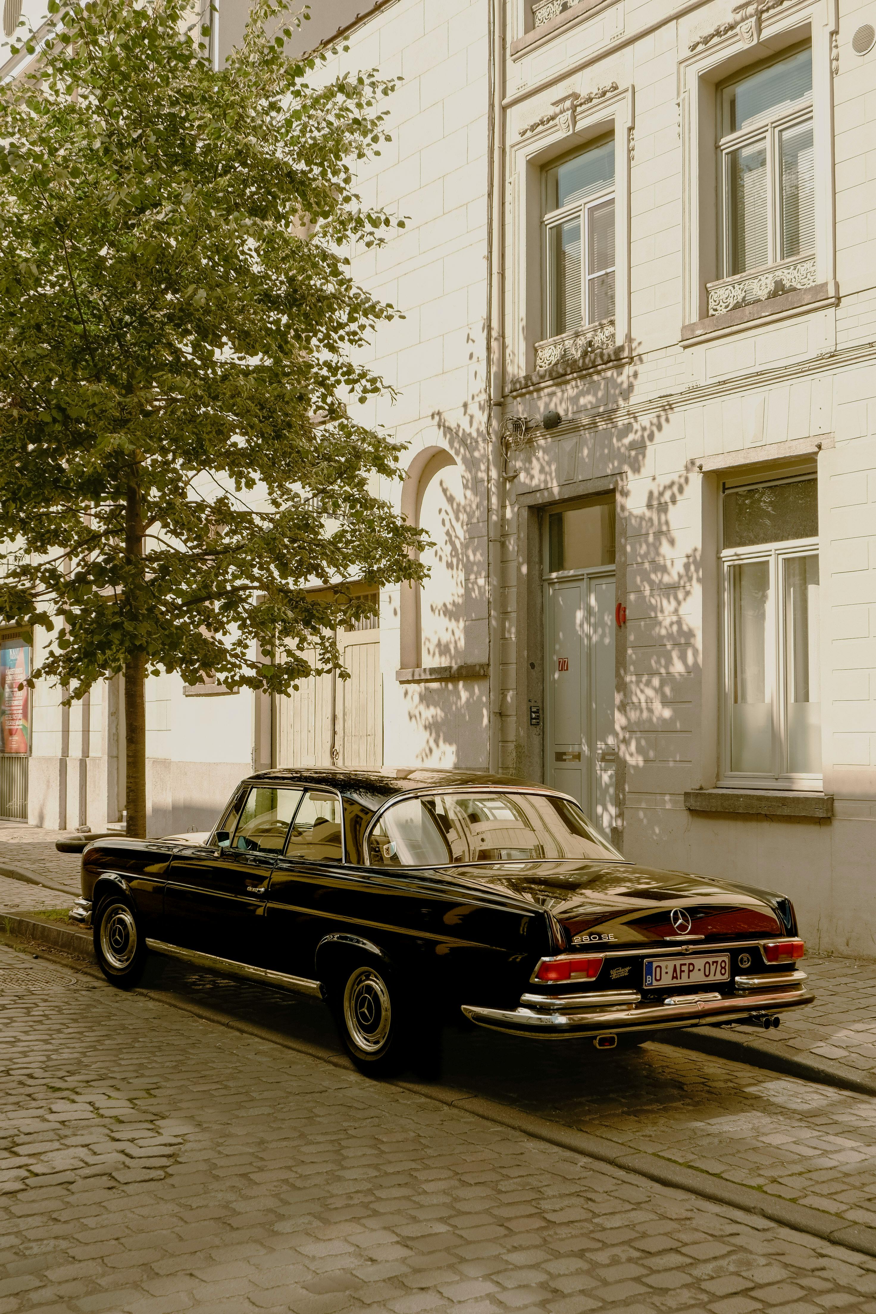 Vintage black Mercedes parked by a tree-lined building on a sunny day.
