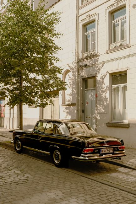 Vintage black Mercedes parked by a tree-lined building on a sunny day.