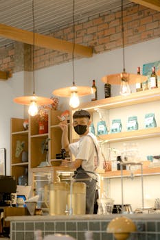 A masked barista prepares drinks in a cozy, warmly lit coffee shop. Ideal for lifestyle and cafe themes.