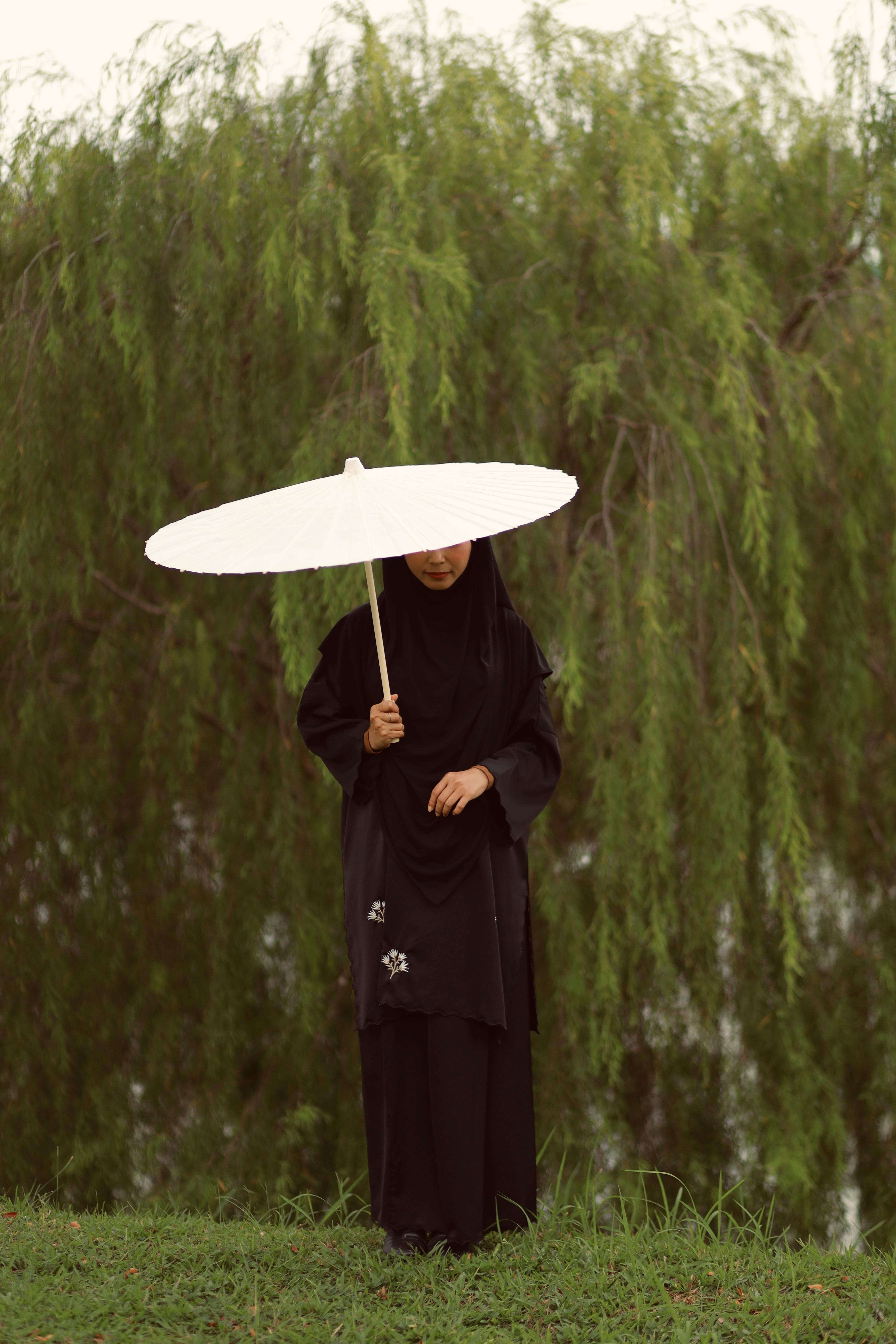A woman stands with a white umbrella beside lush green trees.
