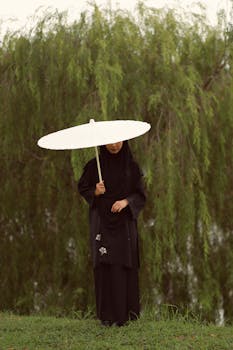 A woman stands with a white umbrella beside lush green trees.