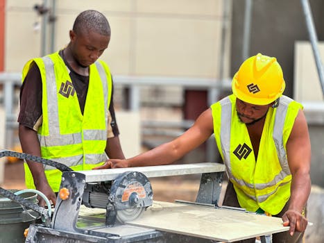 Two construction workers using a tile saw on site, wearing safety gear and high-visibility vests.