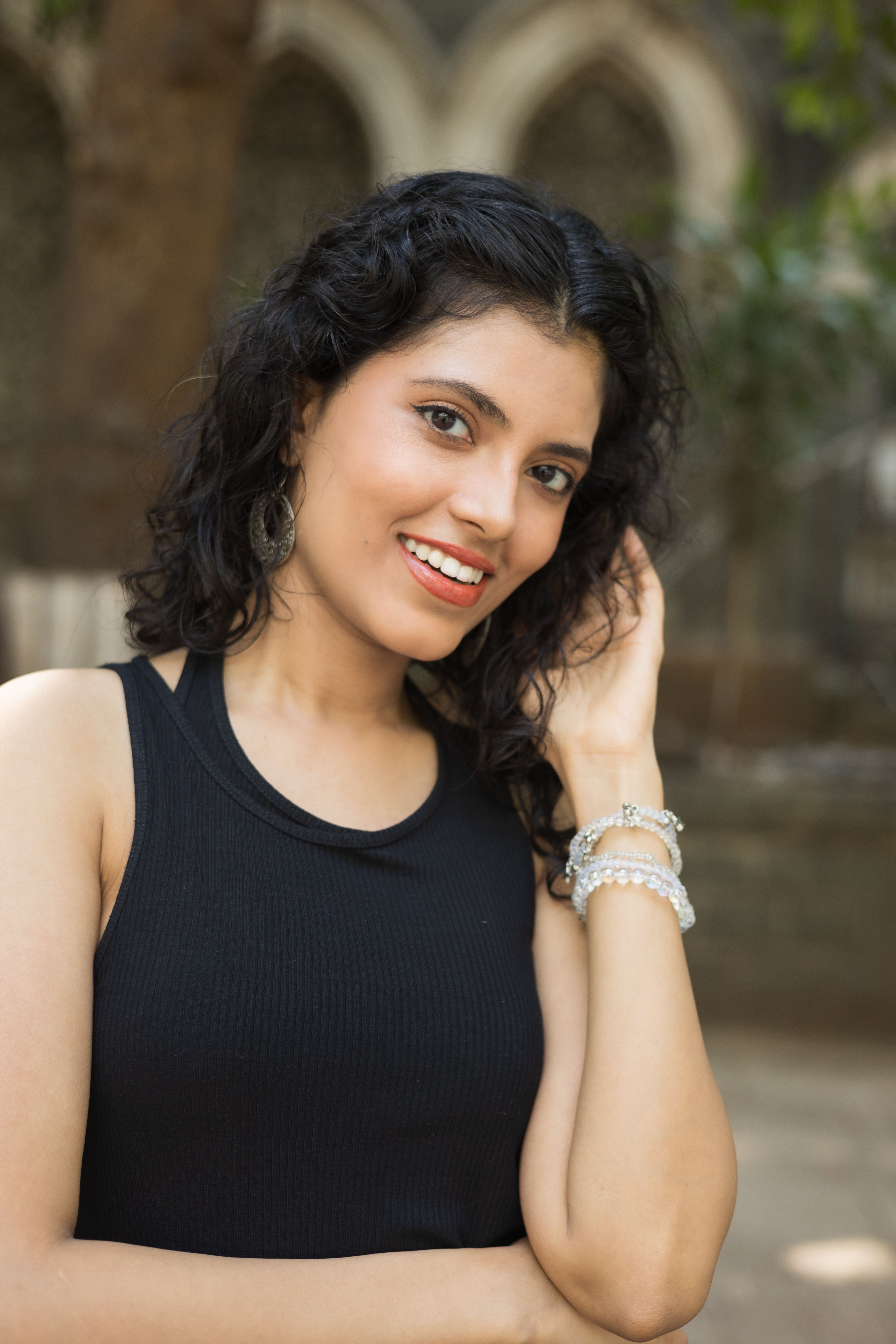 Smiling woman posing outdoors, with natural curly hair and stylish jewelry, against blurred background.