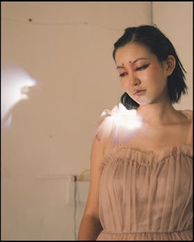 Artistic portrait of a young woman in a delicate dress with creative makeup, glowing in soft indoor lighting.