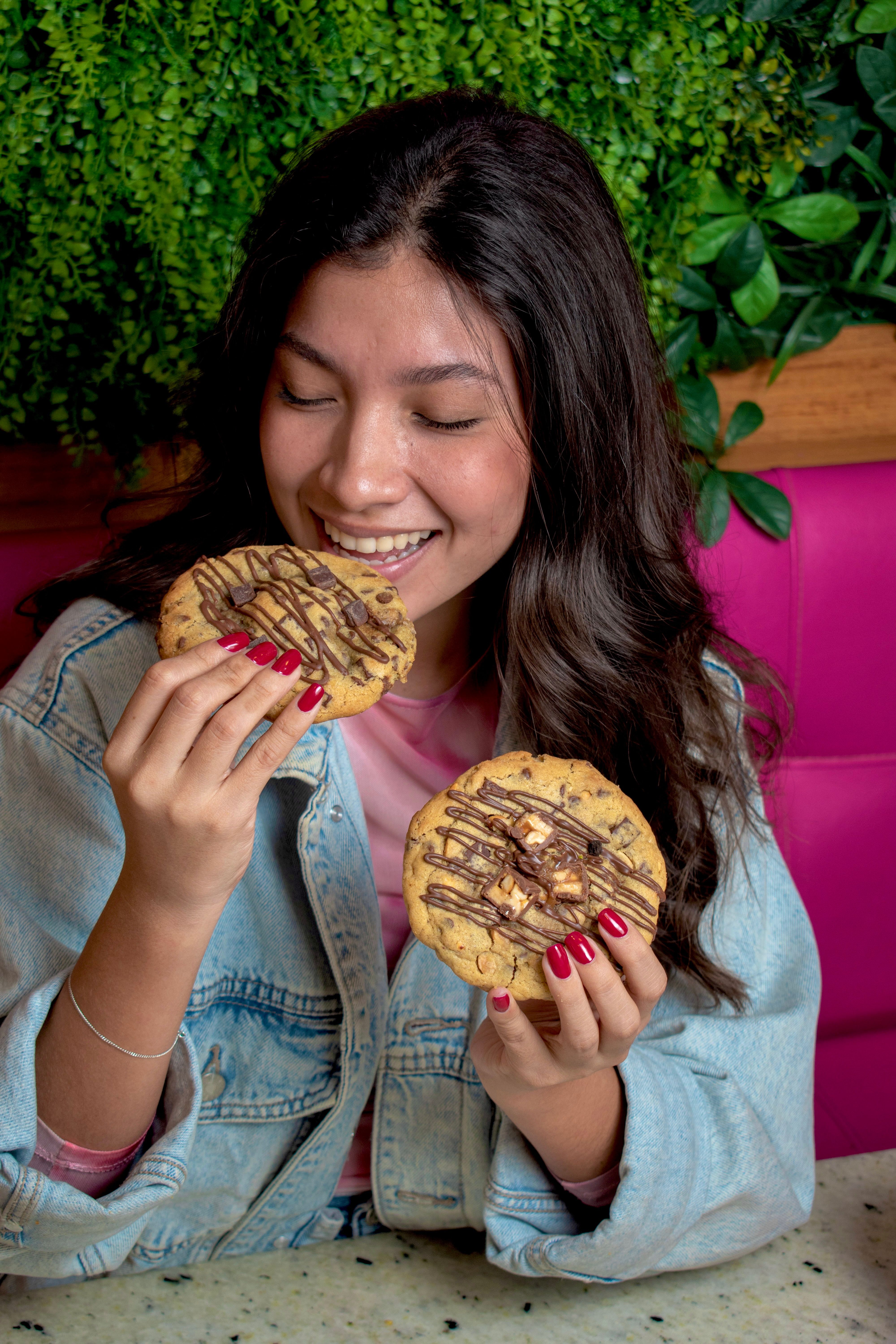 A young woman joyfully savoring two chocolate chip cookies with drizzle indoors.