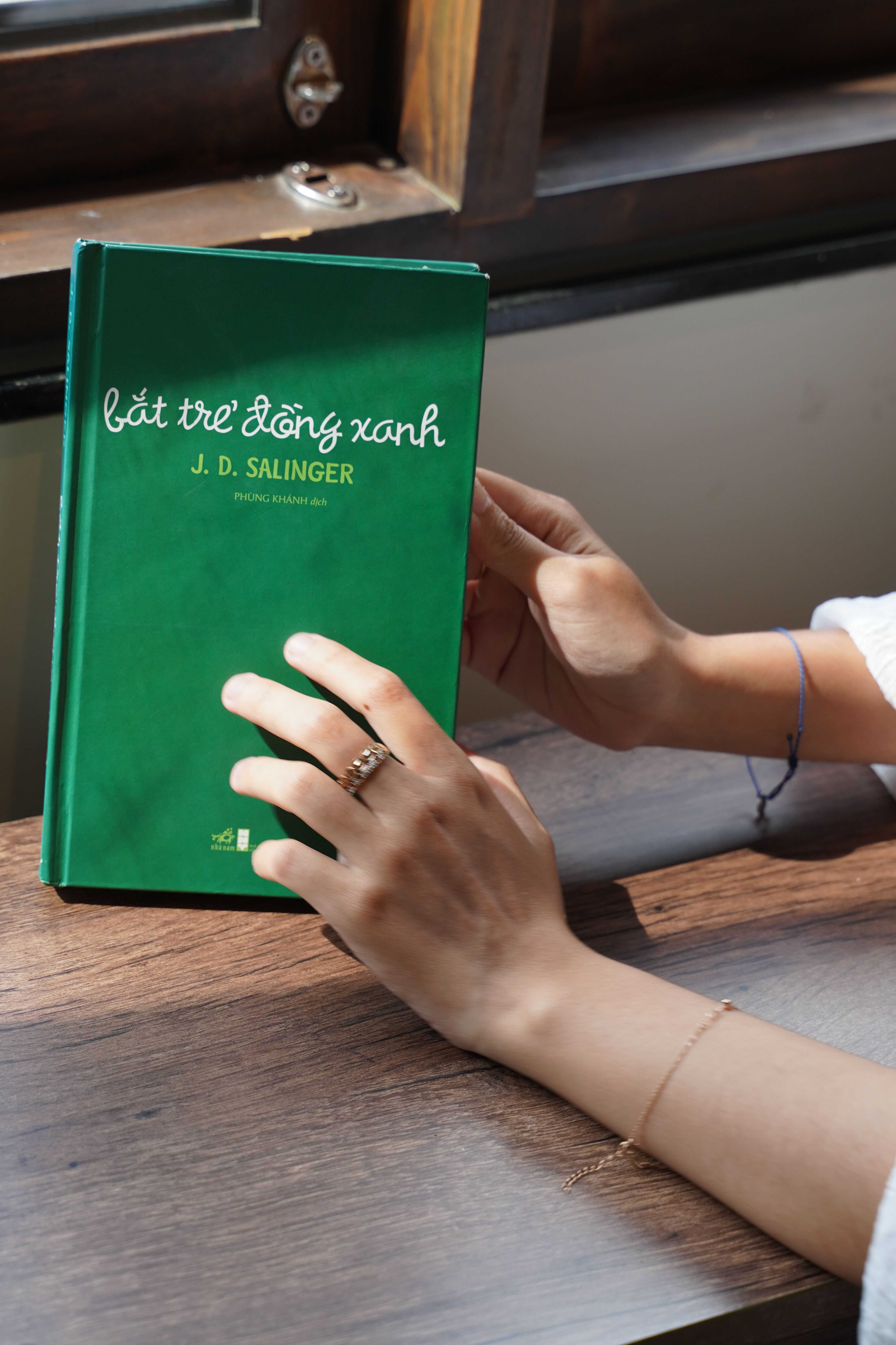 Close-up of hands holding a green book by J.D. Salinger, indoors.