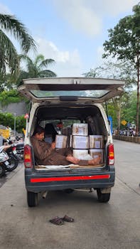 A delivery worker rests in the back of a van surrounded by packages on a city street.