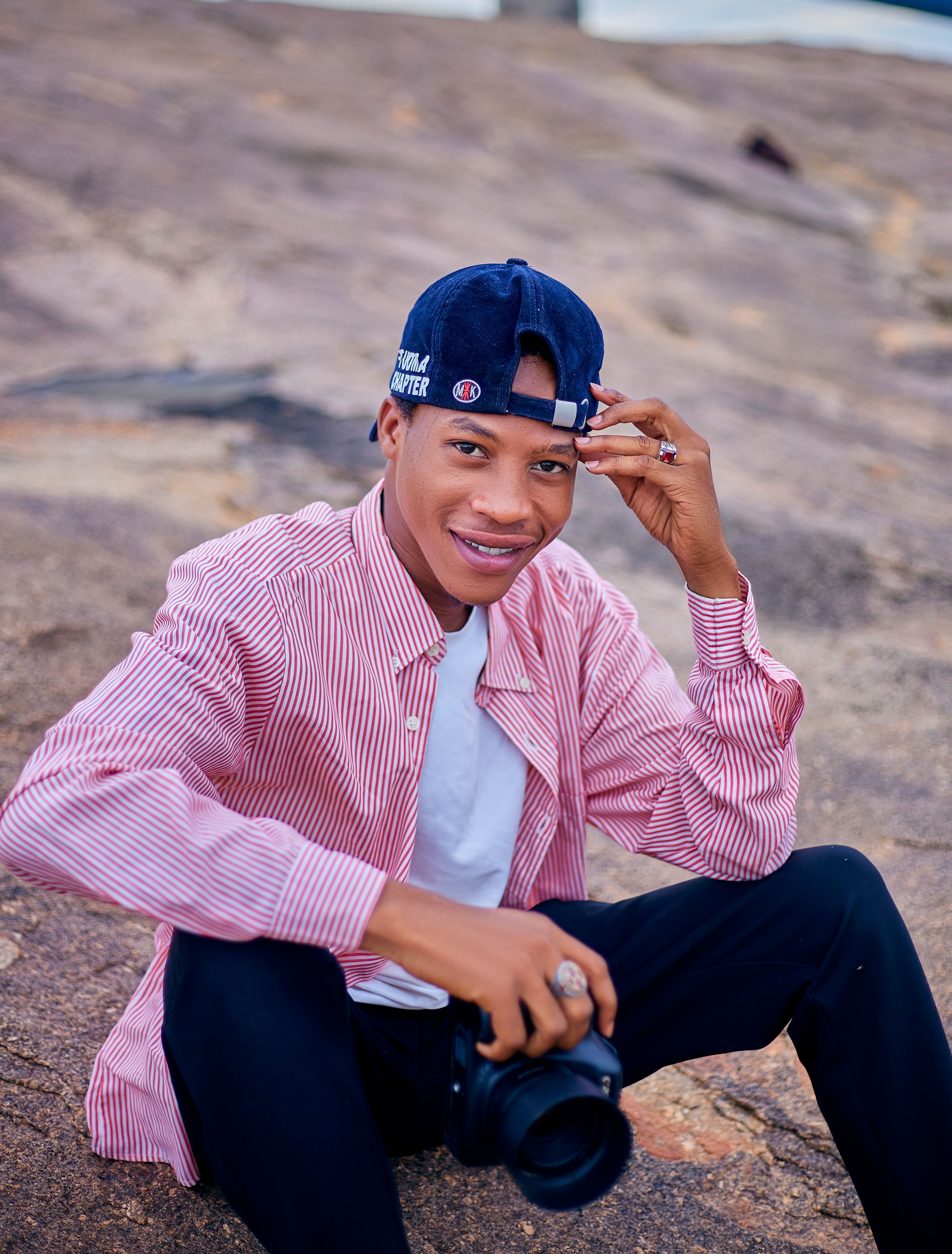Smiling young man wearing a cap, holding a camera outdoors on rocks.