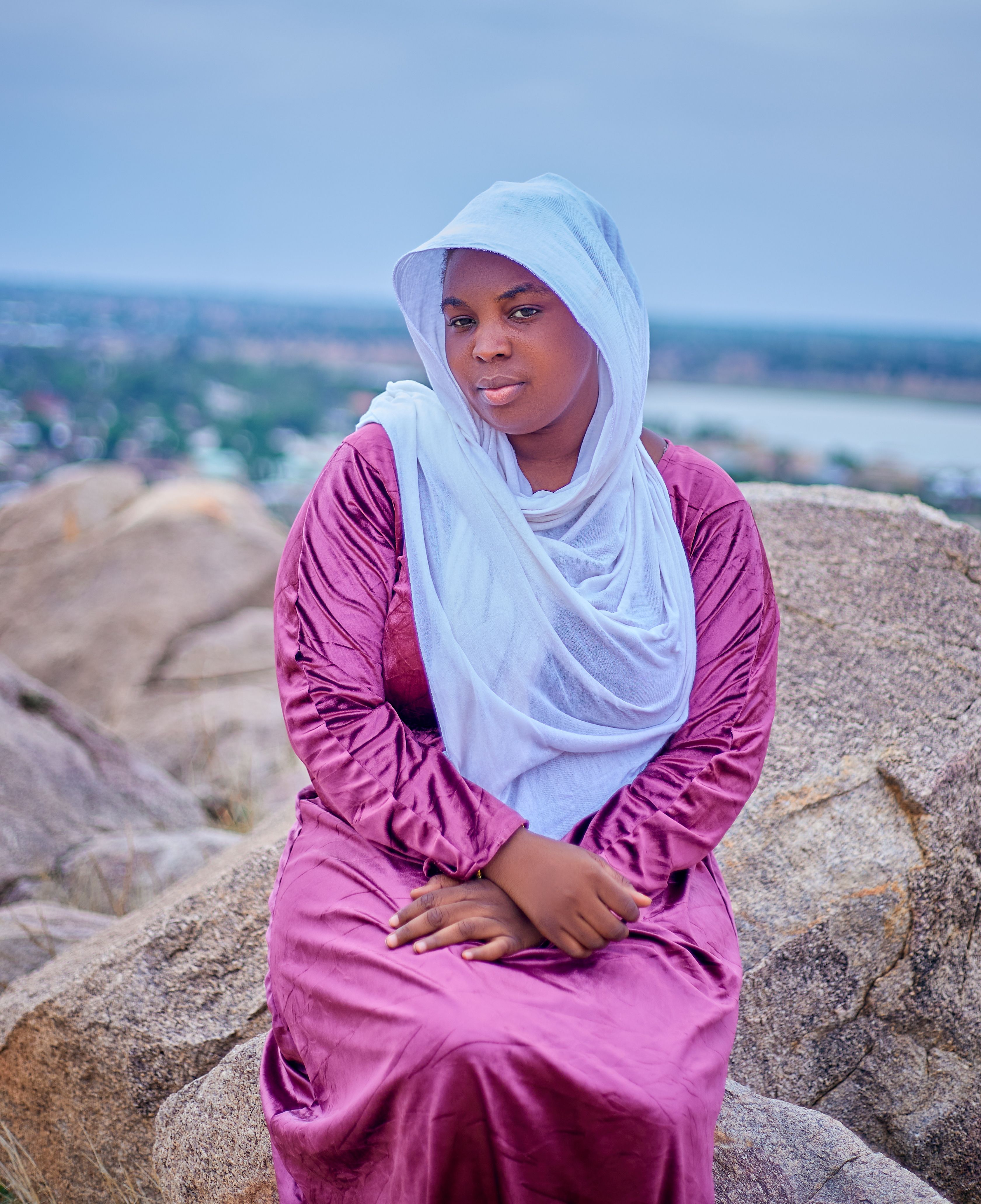 A woman in a purple dress and hijab sits on rocks with a serene backdrop, enjoying the view.