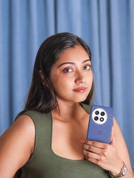 Woman posing confidently with a smartphone against a blue curtain indoors.
