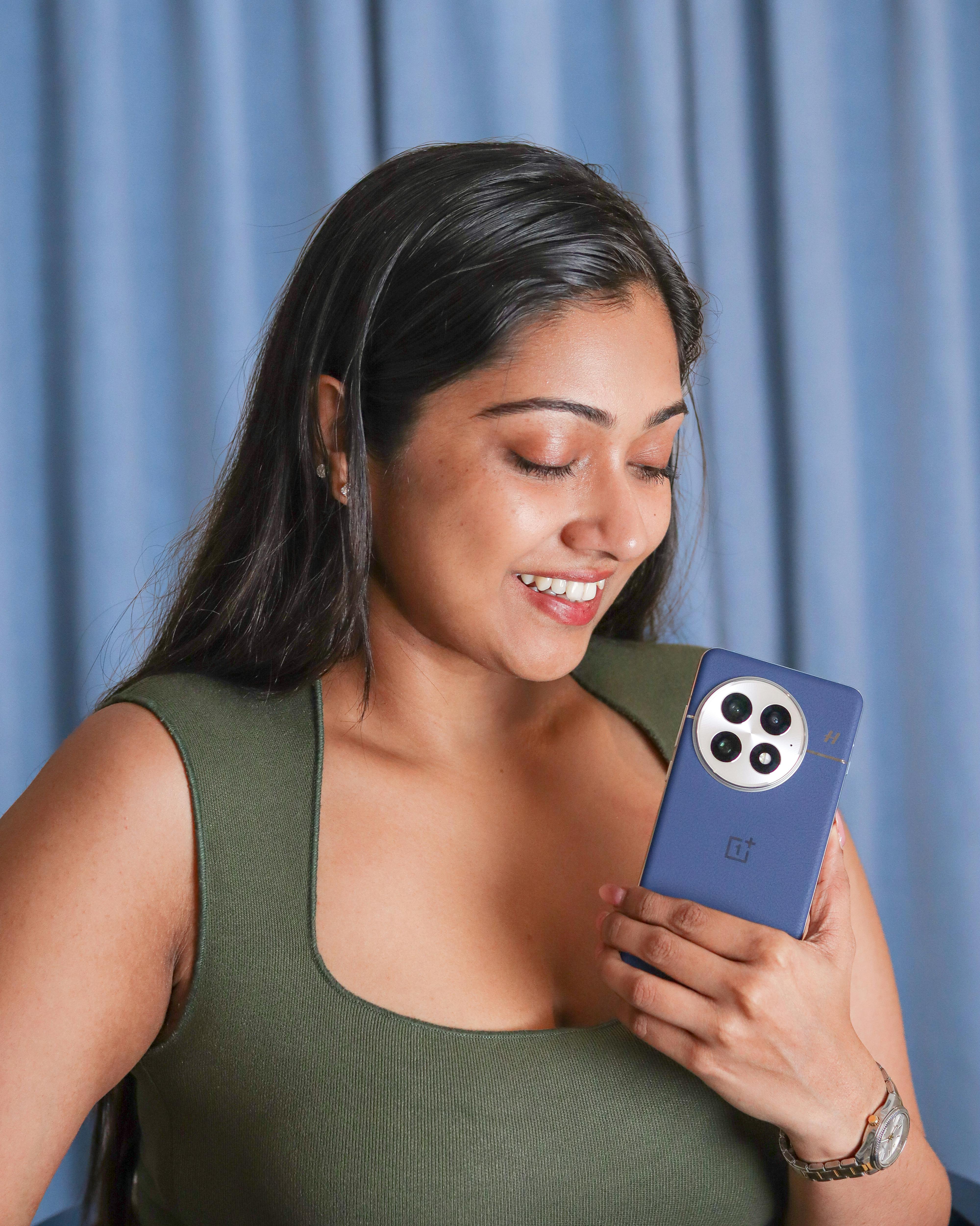 Miễn phí Một người phụ nữ vui vẻ cầm điện thoại thông minh, mỉm cười trên nền rèm màu xanh. Ảnh lưu trữ