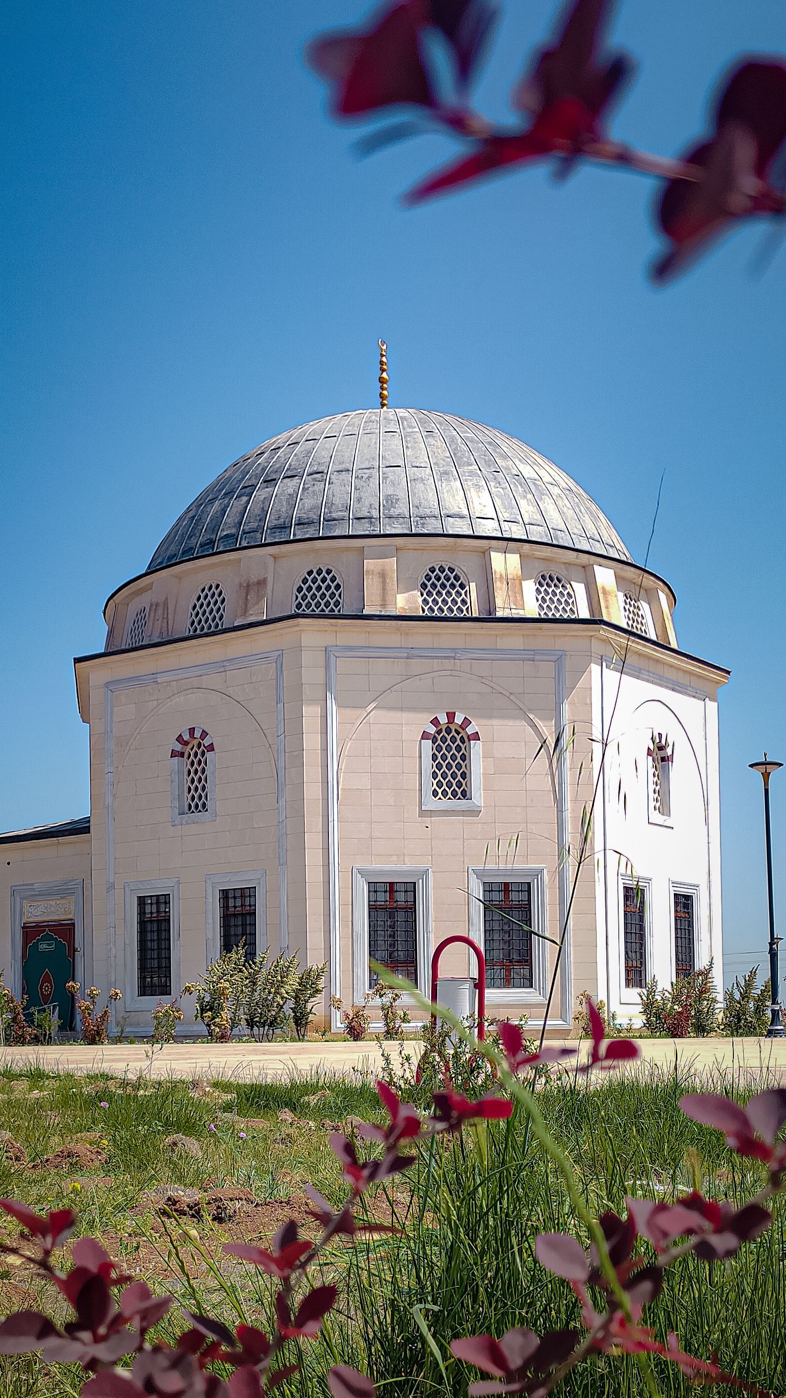 Beautiful Ottoman-style mosque surrounded by greenery and vibrant flowers under a clear blue sky.