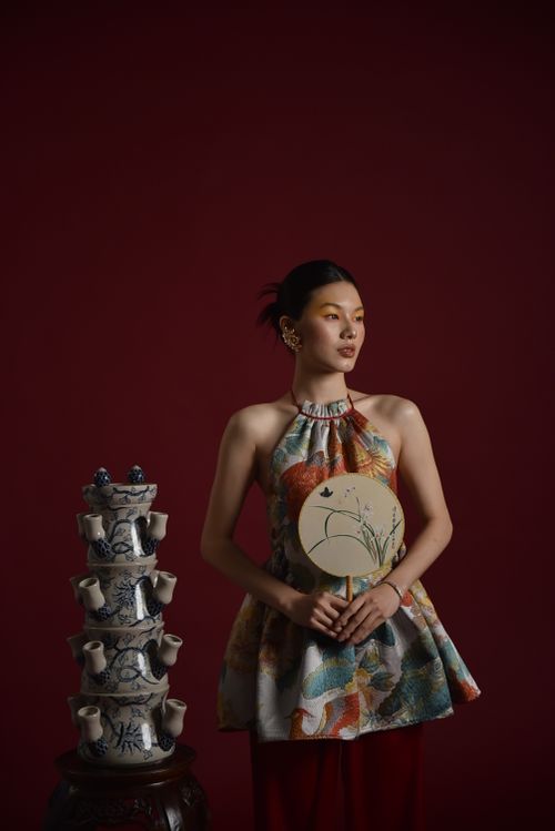 Free Stylish Asian woman holding fan, standing by traditional ceramic teapots against a dark red background. Stock Photo