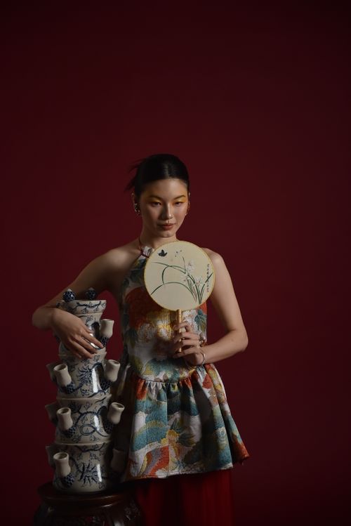 Free Portrait of a woman holding a fan beside a stack of ornate ceramic vases against a red backdrop. Stock Photo