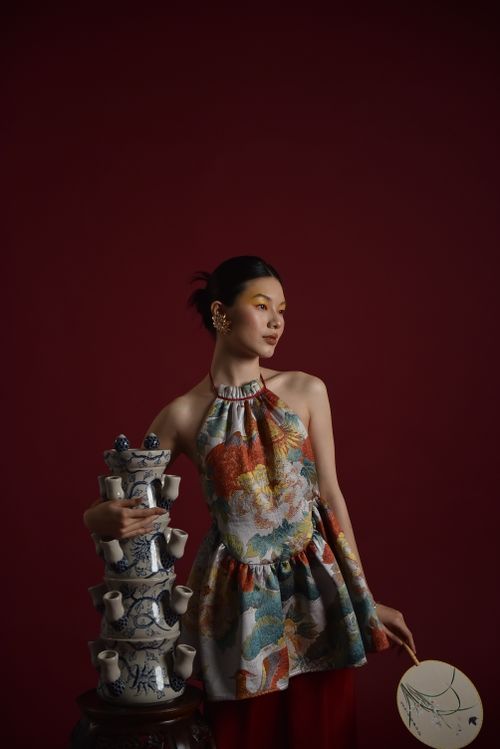 Free Stylish woman in floral dress holding vintage porcelain tower against red backdrop. Stock Photo
