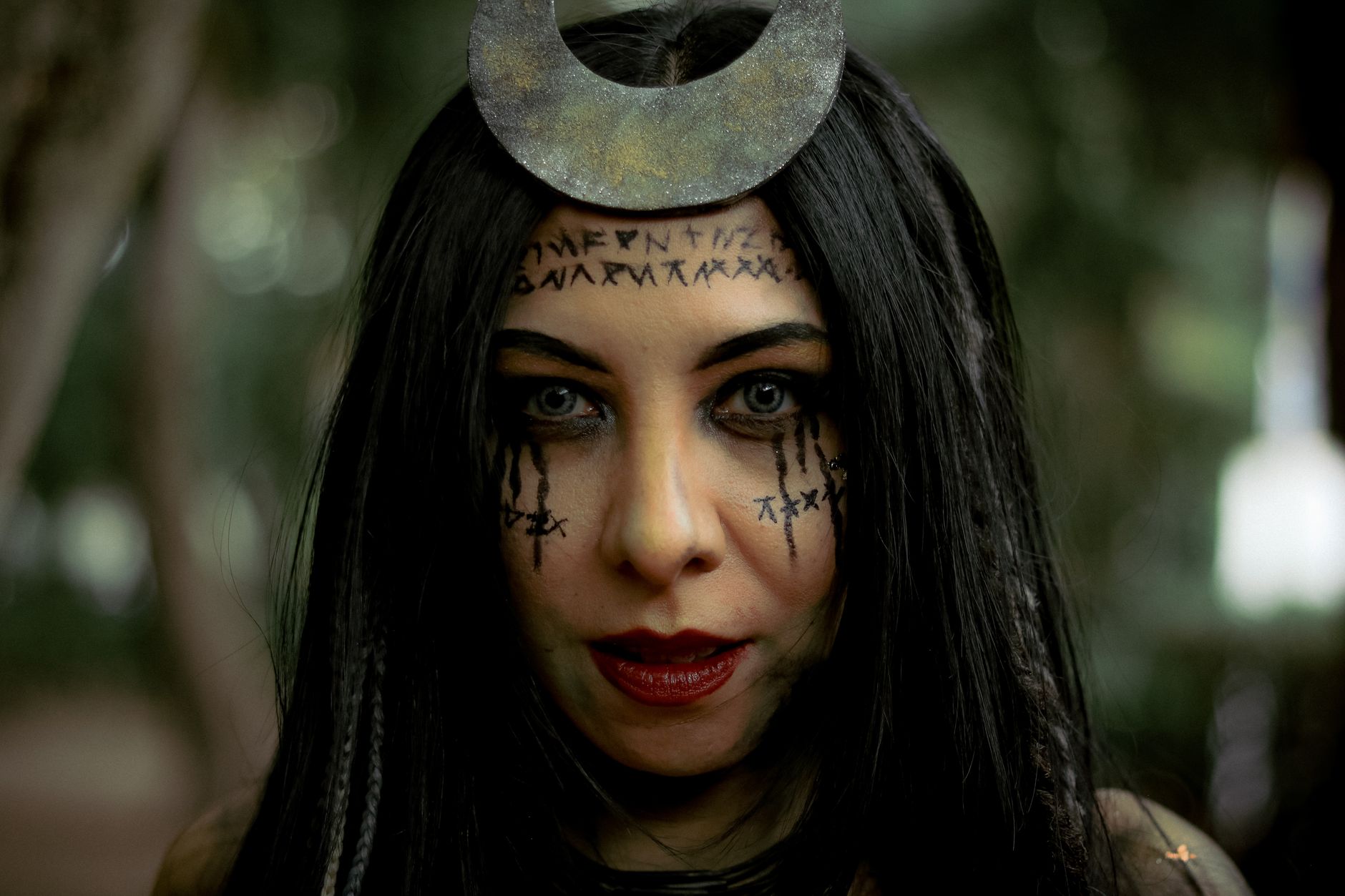 A woman with mystical symbols and dark makeup, set in an outdoor forest environment.