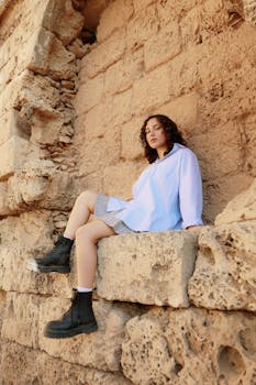 Woman sitting on ancient stone ruins in Cyprus, wearing boots and casual attire, exuding a relaxed aura.