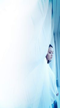 A serene portrait of a woman standing behind a sheer white curtain, gazing out a window.