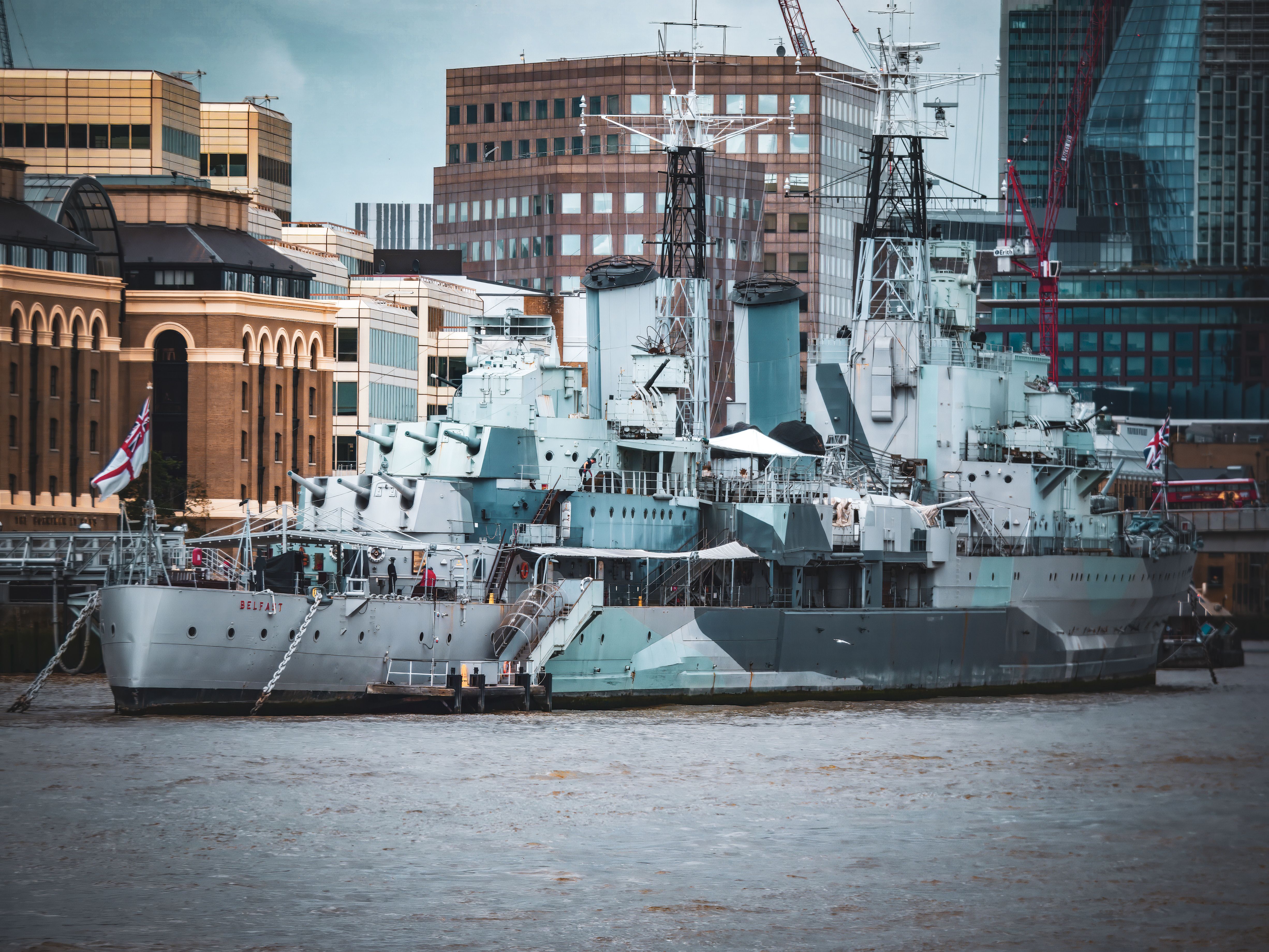 A historic warship docked on a river with a city skyline in the background.