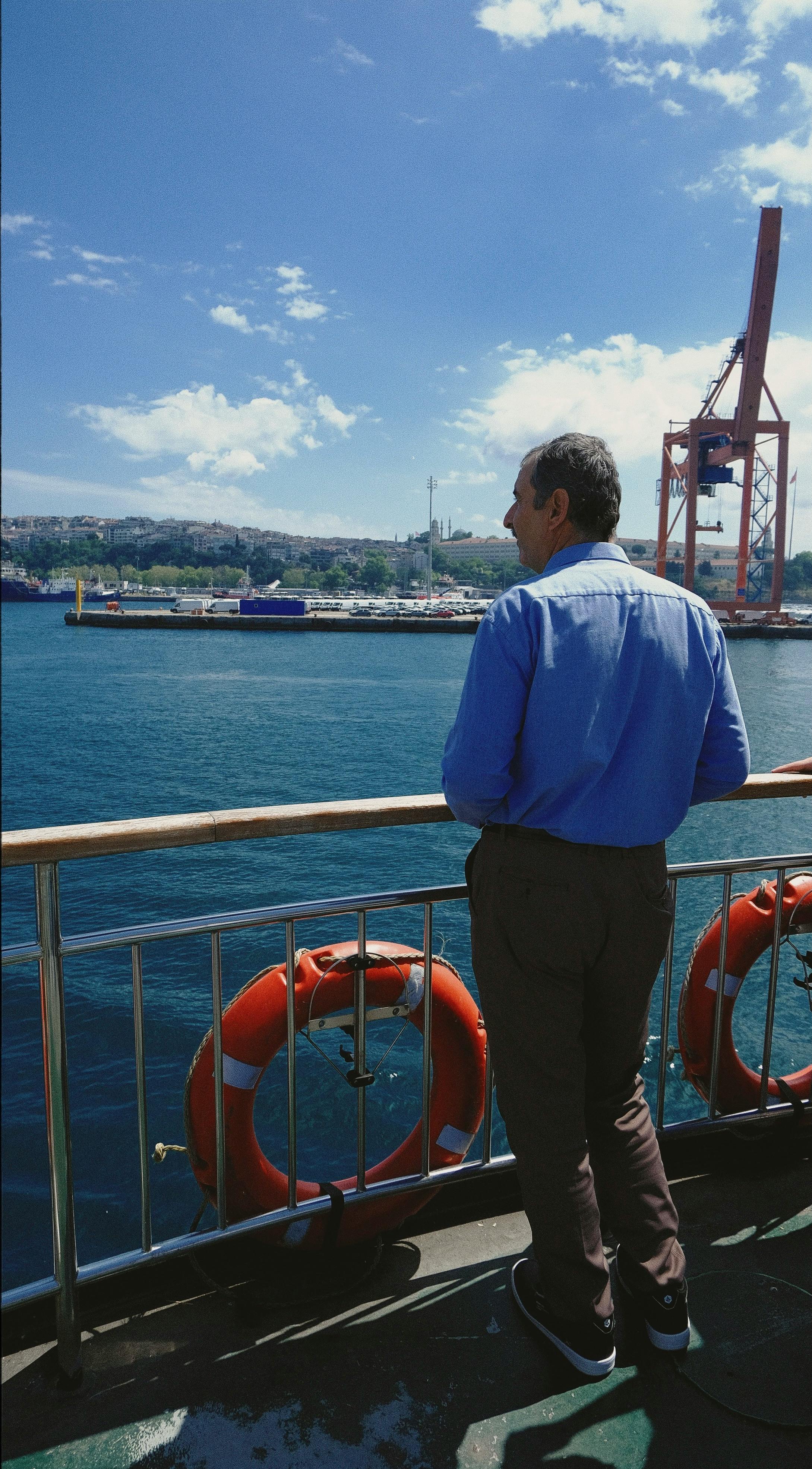 Adult man stands on a ferry deck overlooking a scenic harbor under a clear blue sky.