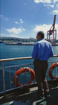Adult man stands on a ferry deck overlooking a scenic harbor under a clear blue sky.