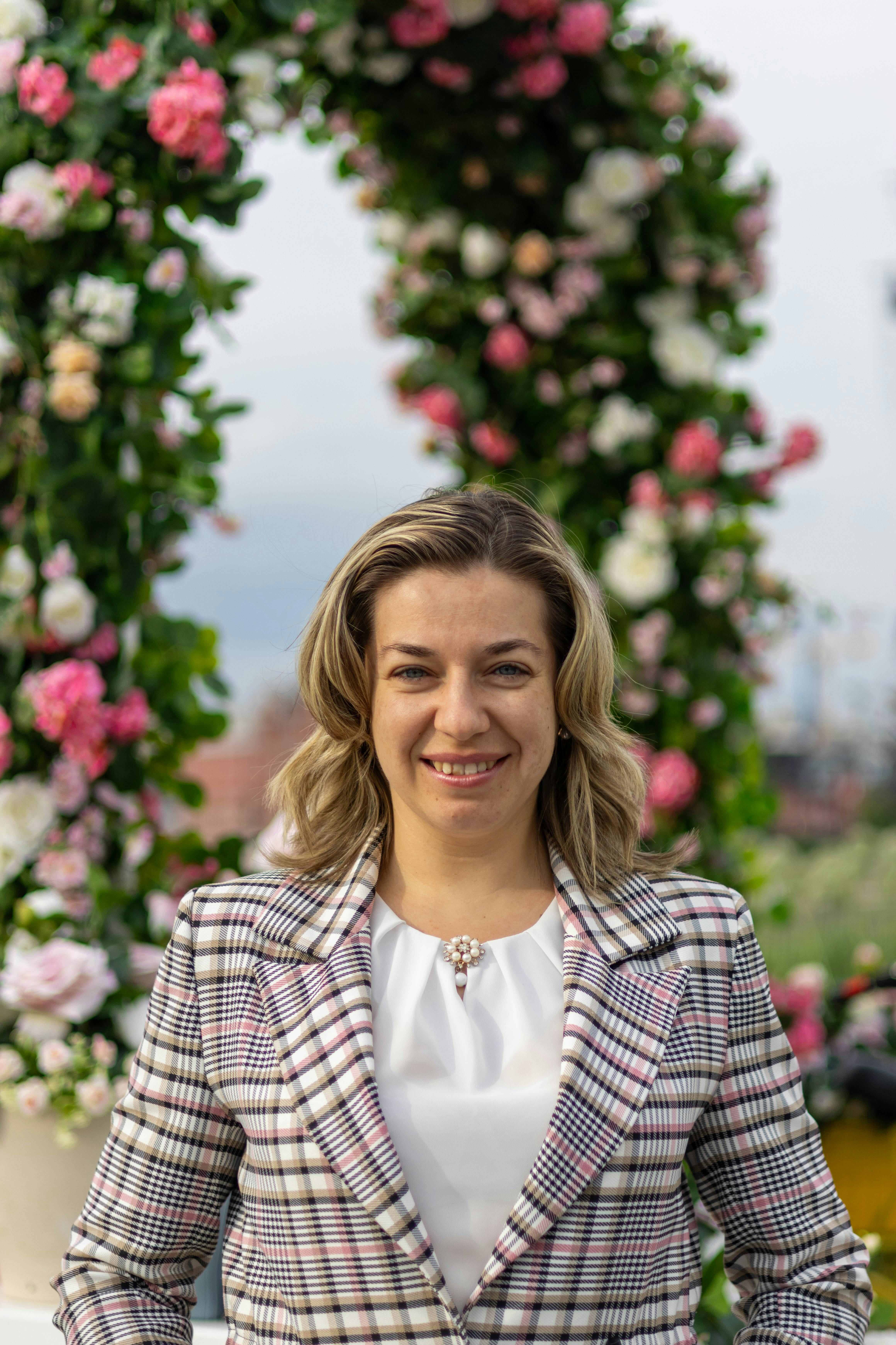 A woman smiling in front of a beautiful flower arch during a daytime outdoor event.