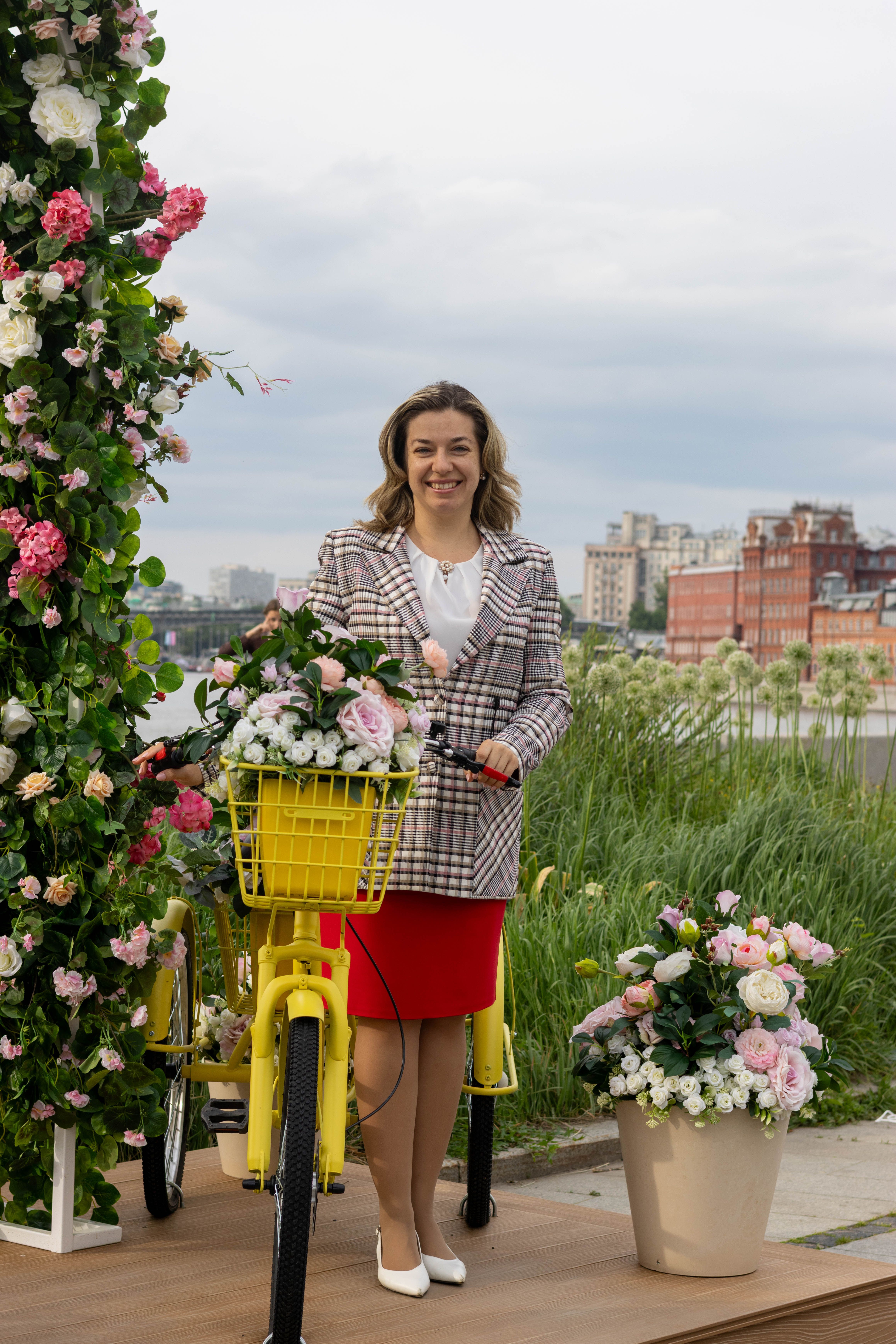Smiling woman with floral arrangements and yellow bike in city park setting.