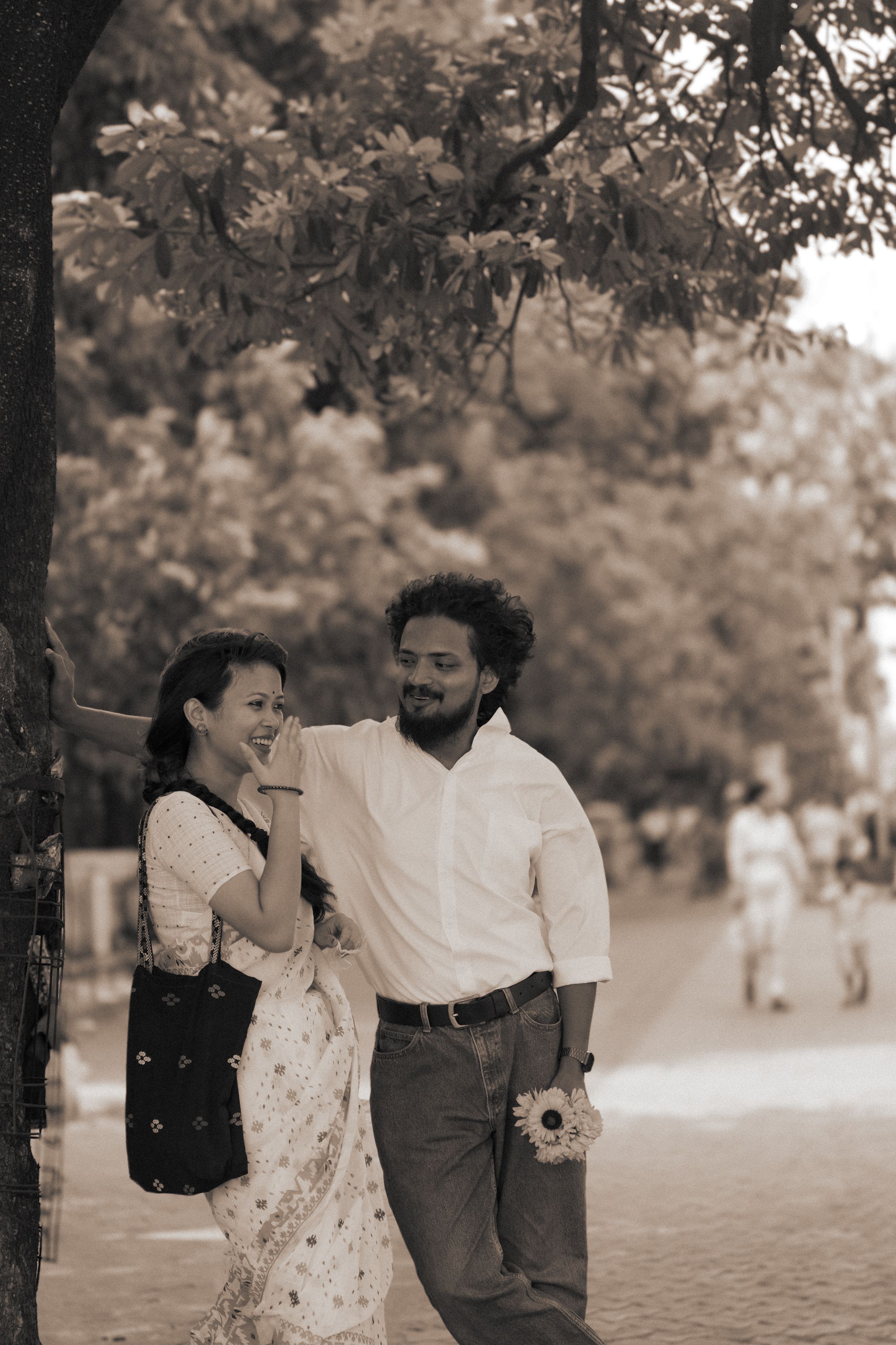 A joyful couple shares a laugh under a tree on a sunny day, holding flowers.