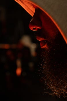 Close-up side profile of a man in a hoodie with a beard, illuminated by dramatic lighting.