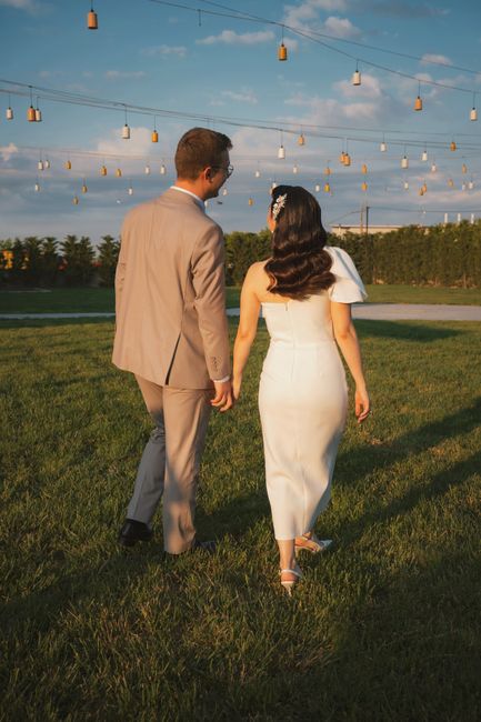A couple in wedding attire walking outdoors under decorative lights at sunset.