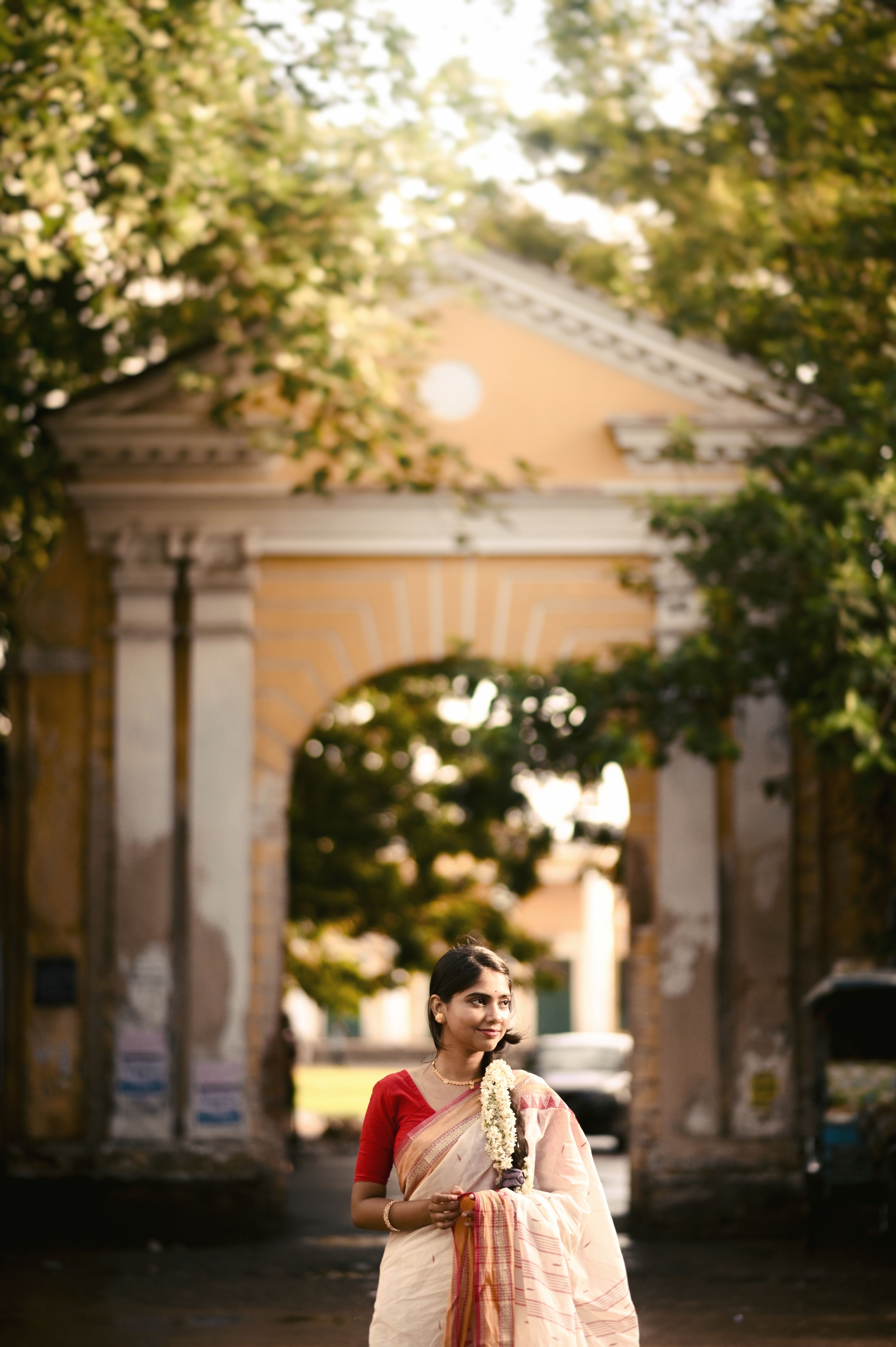 A woman in traditional Indian attire stands gracefully near an archway, surrounded by greenery.