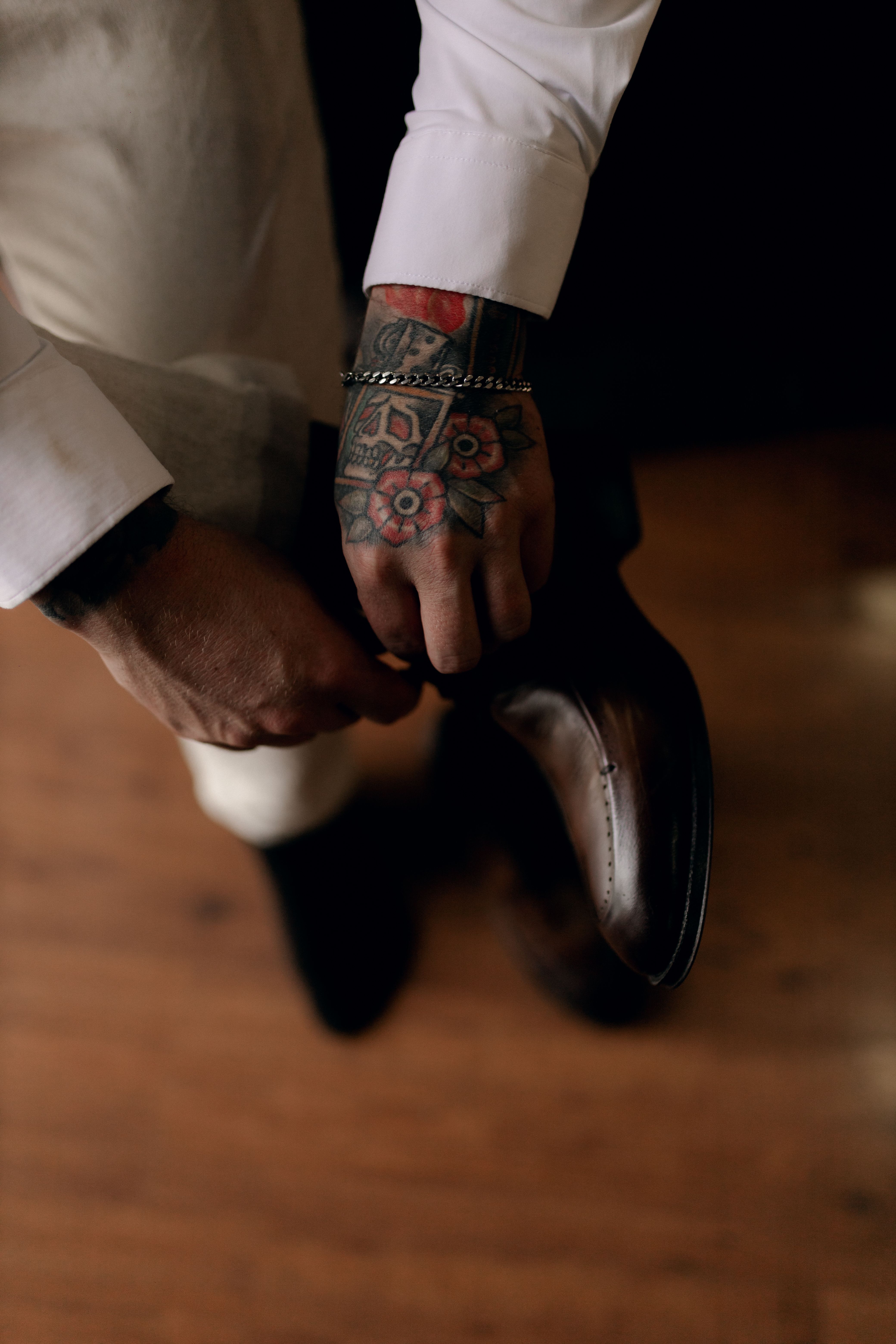 A man with tattoos on his arm adjusts a formal leather shoe indoors.