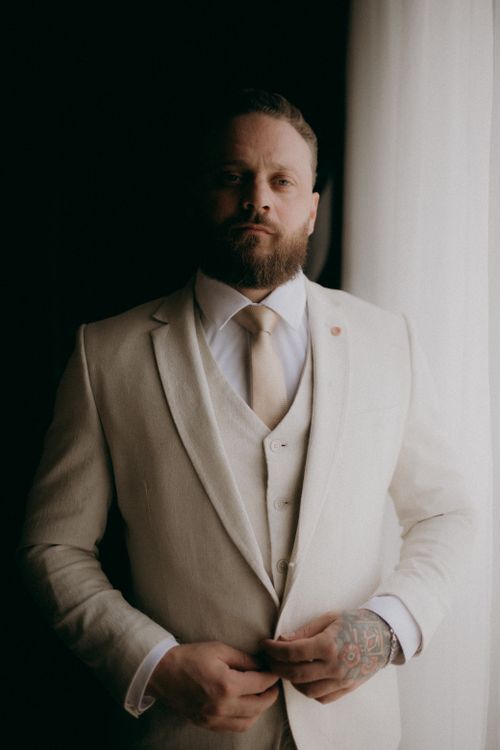Free Elegant portrait of a bearded man standing in a beige suit near a window with soft lighting. Stock Photo