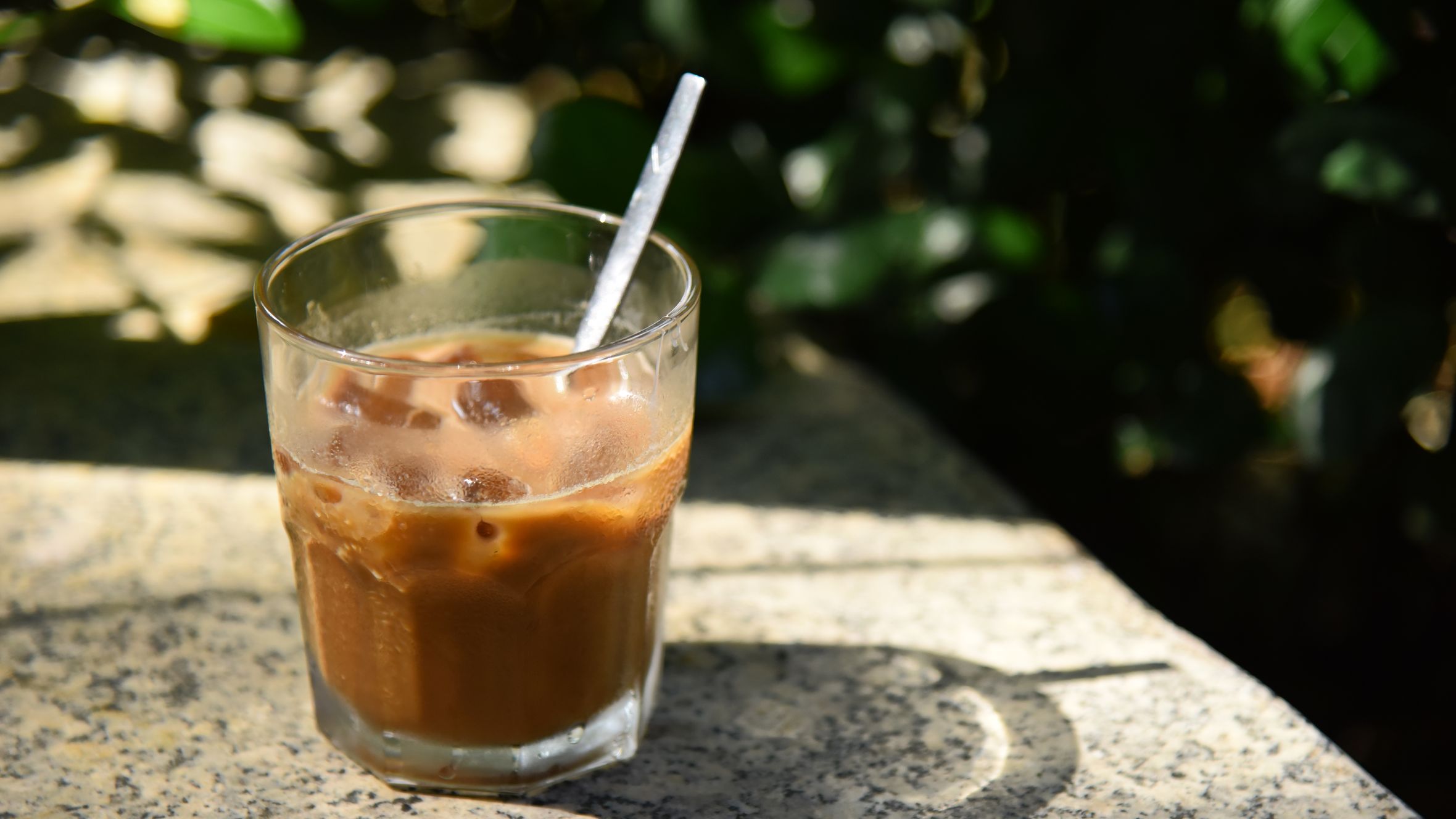 A cool glass of iced coffee on a sunny outdoor granite table, surrounded by greenery.