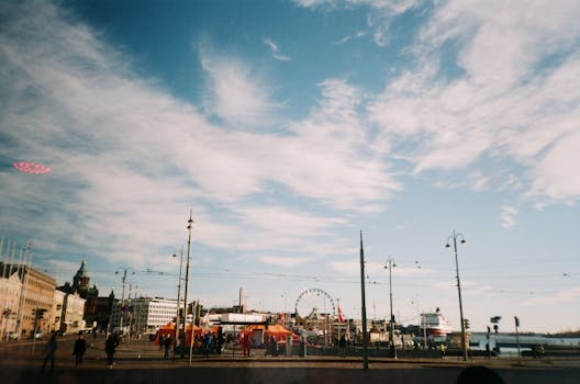 A ferris wheel in the Helsinki cityscape against a blue and cloudy sky. by Inga Seliverstova