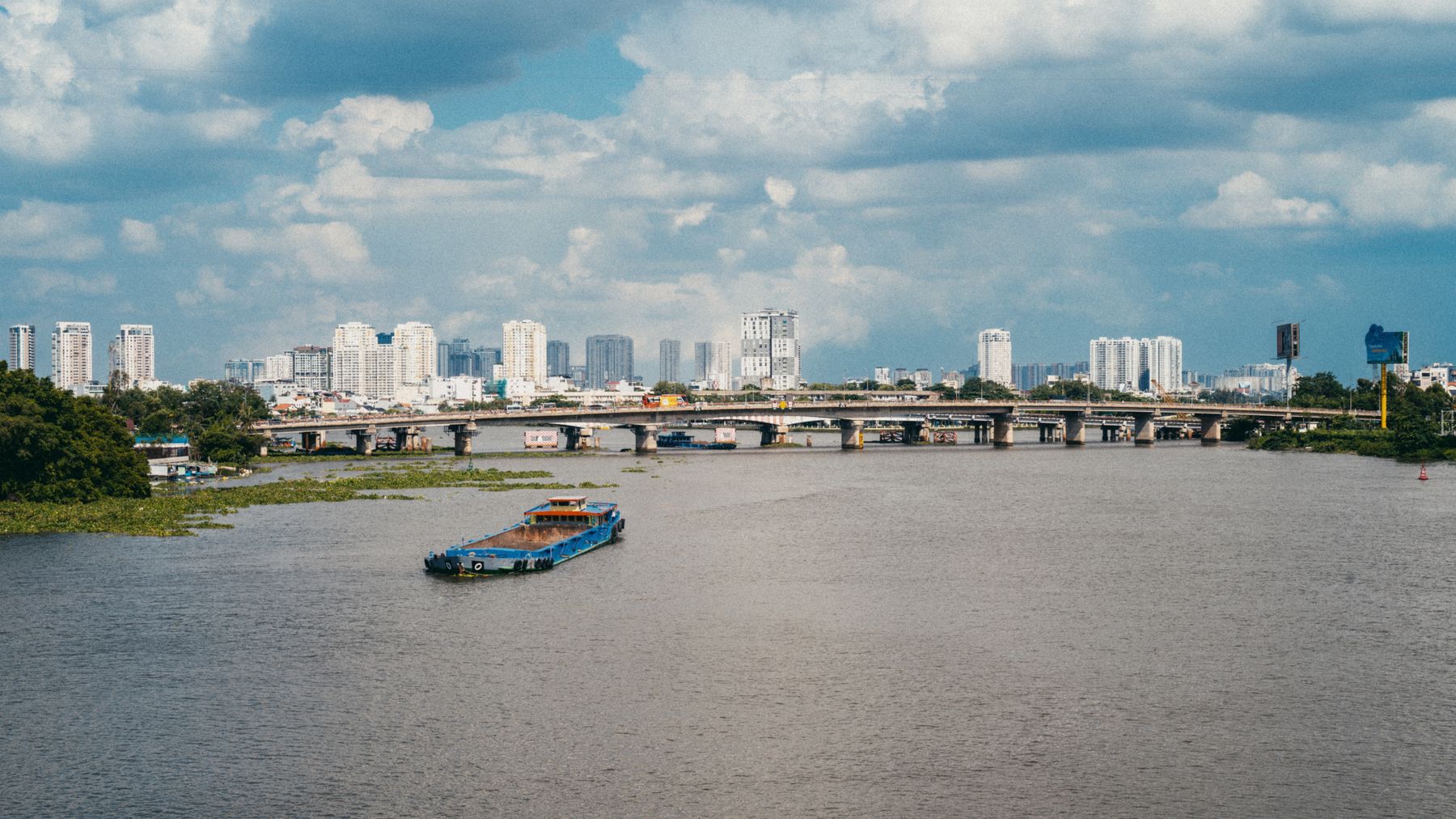 A bustling view of the Saigon River with a barge and the city skyline under a blue sky.
