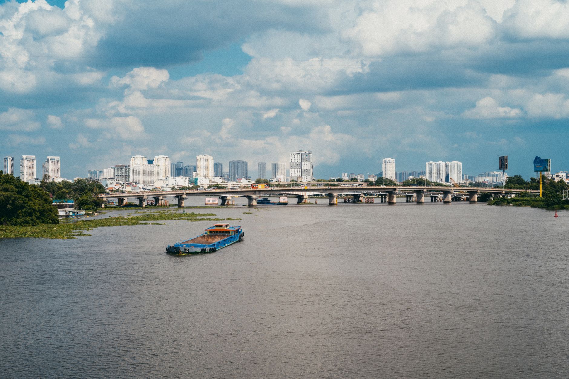 A bustling view of the Saigon River with a barge and the city skyline under a blue sky.