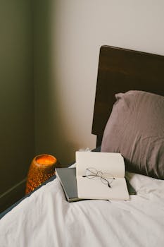 A cozy bedroom scene with open book, glasses, and warm lamp on bedside table.