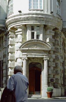 An older adult walks past the grand entrance of a historic stone building in daylight.