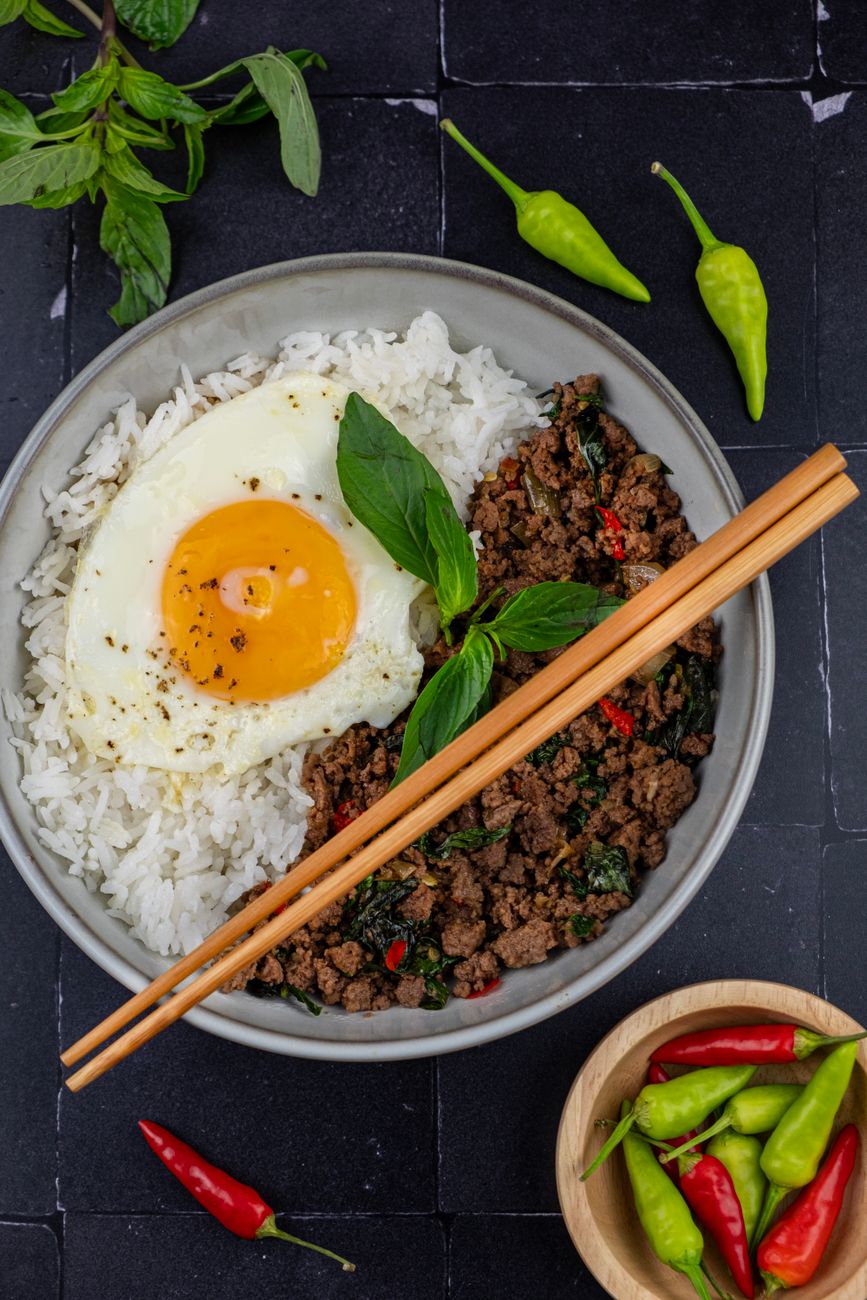 White rice bowl topped with dark stir-fried ground pork mixed with wilted green holy basil leaves and crowned with a golden fried egg with crispy edges
