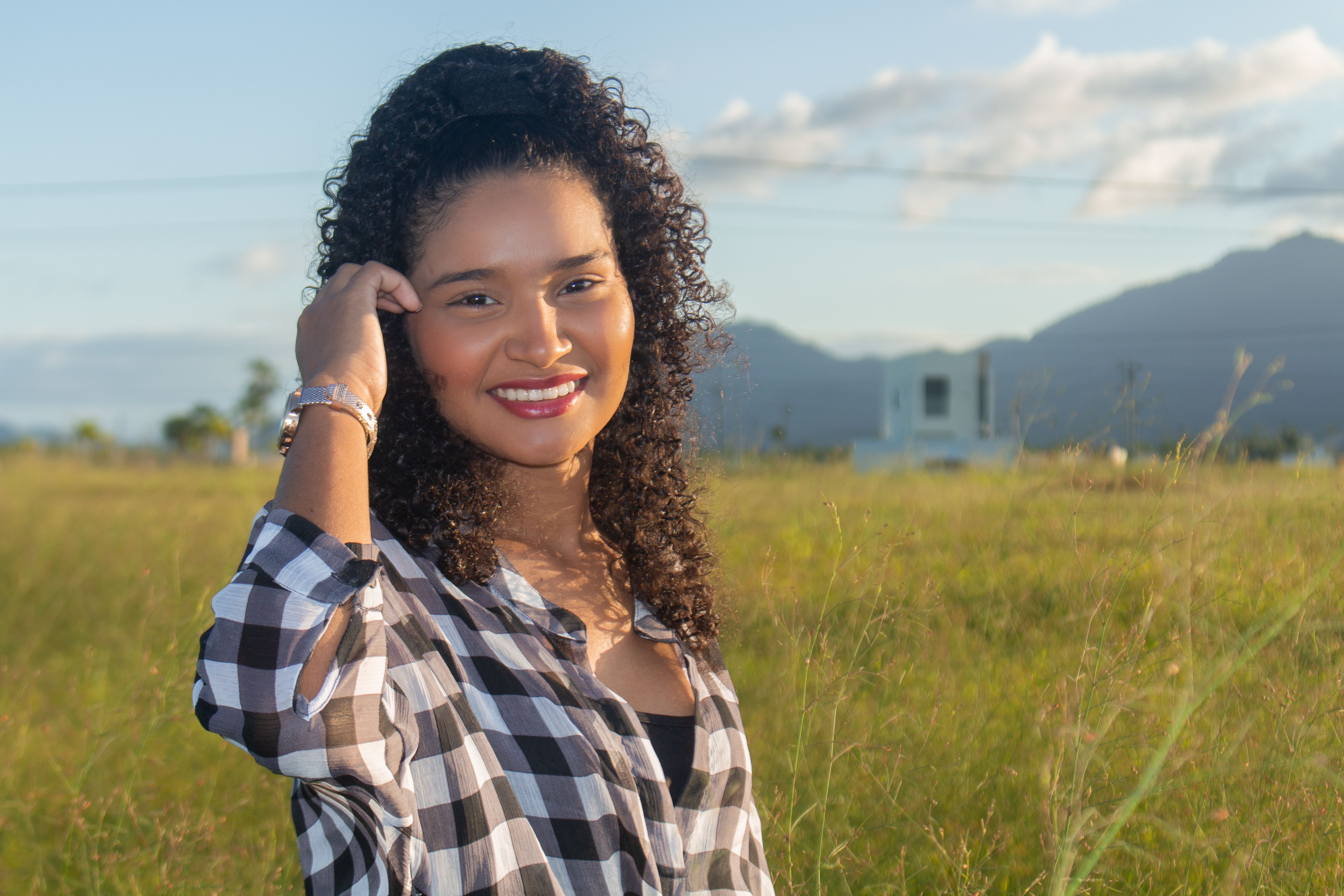 A young woman with curly hair smiling in a grassy field on a sunny day.
