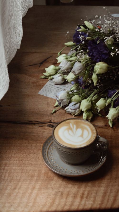 Free A cozy scene featuring a cappuccino with latte art on a rustic table alongside a floral bouquet. Stock Photo