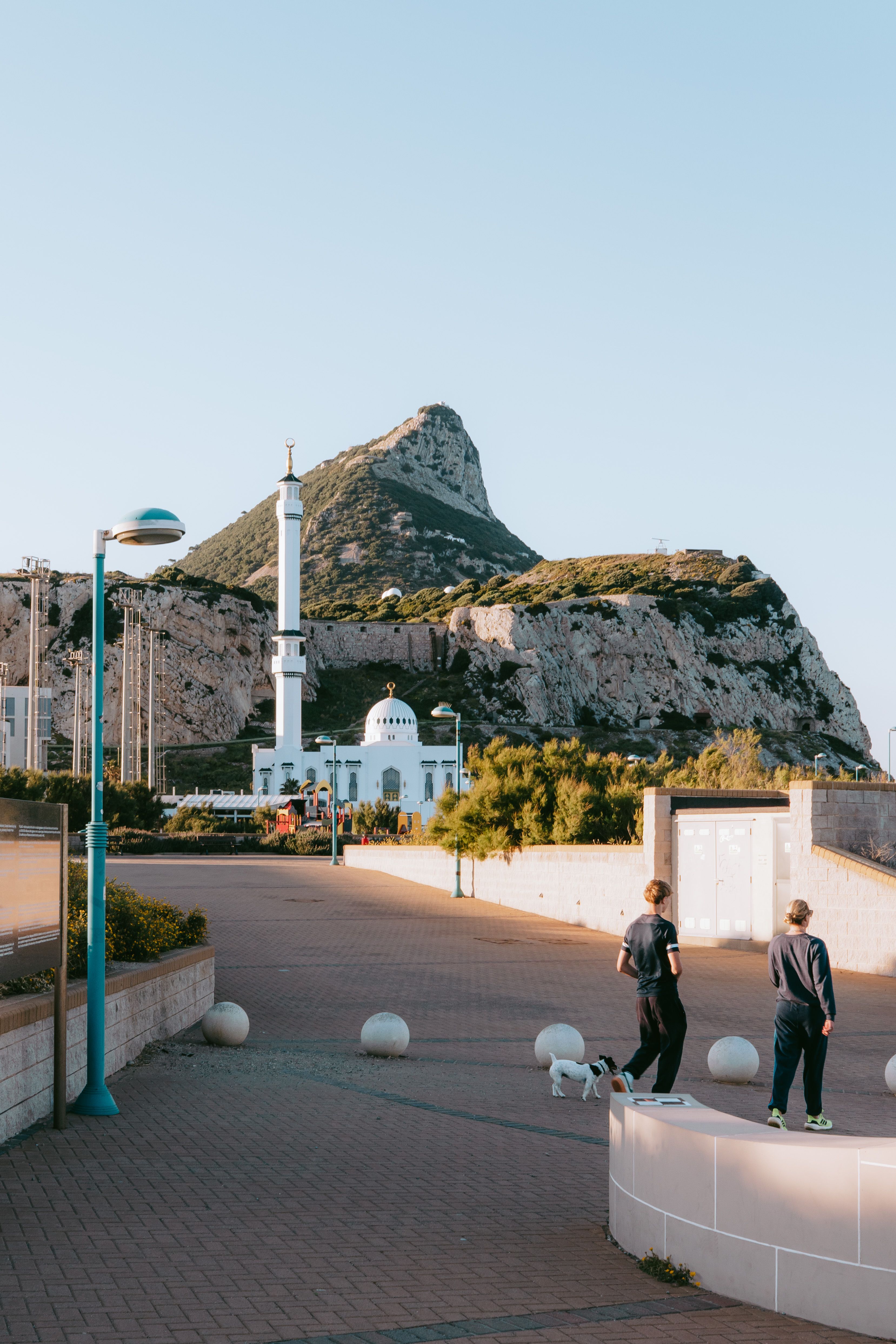 Couple walking dog near Ibrahim-al-Ibrahim Mosque in Gibraltar during golden hour with Mediterranean coast and mountains in view.