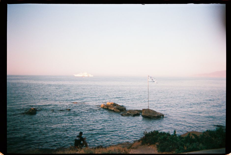 A peaceful scene by the Greek sea featuring a flag and distant cruise ship under a clear sky.